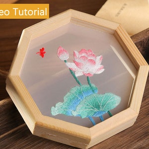 May include: A wooden octagonal frame with a white background and an embroidered image of a pink and white lotus flower with green leaves. The frame is on a wooden surface.