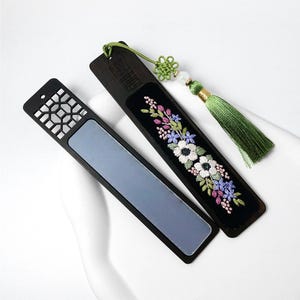 Peut inclure: Deux marque-pages rectangulaires en bois sombre. L'un présente un motif géométrique découpé sur le haut et un centre gris lisse. L'autre possède une incrustation de velours noir avec une broderie florale blanche, violette et rose, ornée d'un pompon vert.