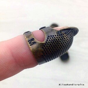 May include: A brass thimble with a textured surface and a hole for the finger. The thimble is marked with the letter "M".
