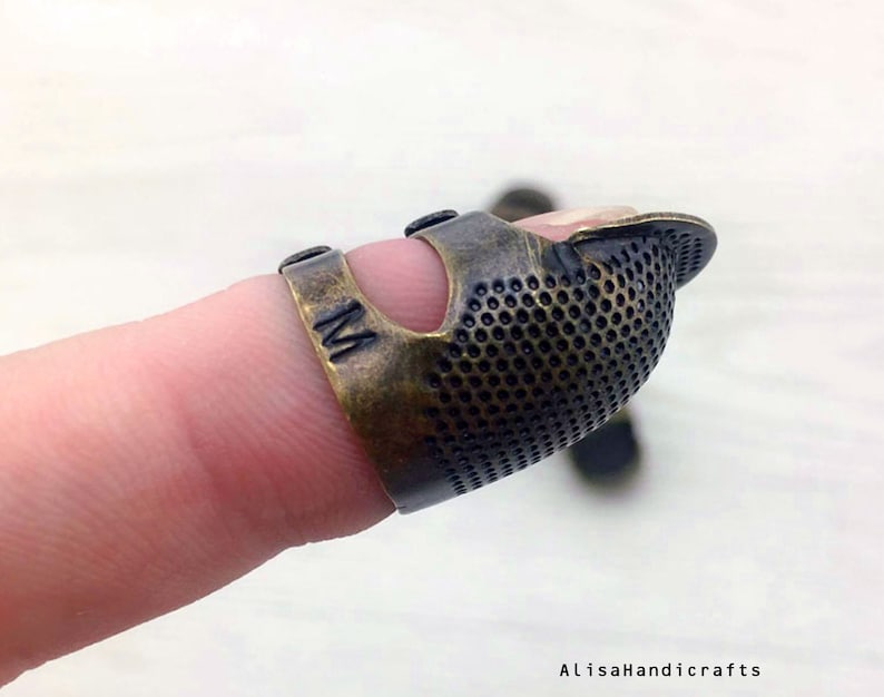 May include: A brass thimble with a textured surface and a hole for the finger. The thimble is marked with the letter "M".