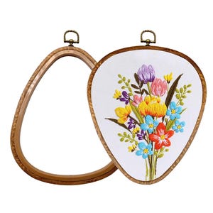 May include: Two teardrop-shaped embroidery hoops with a wood-grain finish and a brass-colored hanging loop at the top. One hoop is empty, while the other contains white fabric featuring a colorful embroidered bouquet of tulips and wildflowers.