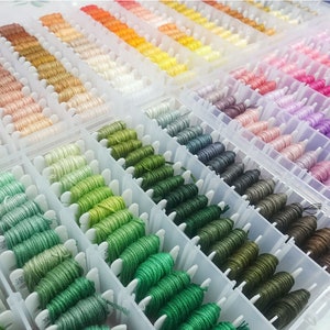 May include: A clear plastic storage box filled with rows of colorful embroidery floss spools. The spools are organized by color, with shades of green, blue, purple, pink, orange, and yellow.