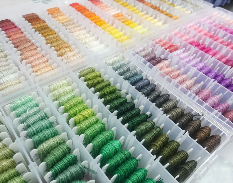 May include: A clear plastic storage box filled with rows of colorful embroidery floss spools. The spools are organized by color, with shades of green, blue, purple, pink, orange, and yellow.