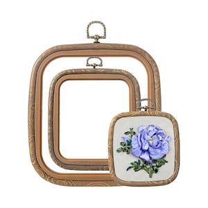May include: Two empty square embroidery hoops with a wood-grain texture and bronze-colored hanging hardware, nested inside each other, alongside a smaller square embroidery hoop containing a purple ribbon-embroidered flower on white fabric.