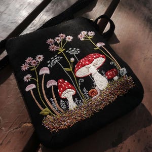May include: A black crossbody bag featuring detailed embroidery of red and white mushrooms, small pink wildflowers, and green foliage. The design includes a textured ground layer of brown and green thread, with a black strap attached to the top.