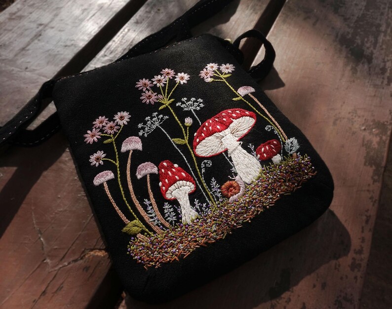May include: A black crossbody bag featuring detailed embroidery of red and white mushrooms, small pink wildflowers, and green foliage. The design includes a textured ground layer of brown and green thread, with a black strap attached to the top.