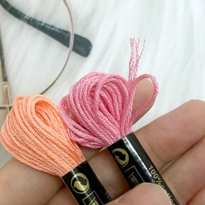 May include: Two spools of embroidery floss, one pink and one peach, with black labels that say "100% Cotton".