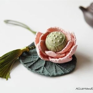 May include: A pink and green fabric lotus flower charm with a green tassel. The flower is on a green fabric leaf with a textured design.