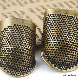 May include: Two antique gold-toned thimbles with a textured, dotted surface. The thimbles are made of metal and have a slightly rounded shape.