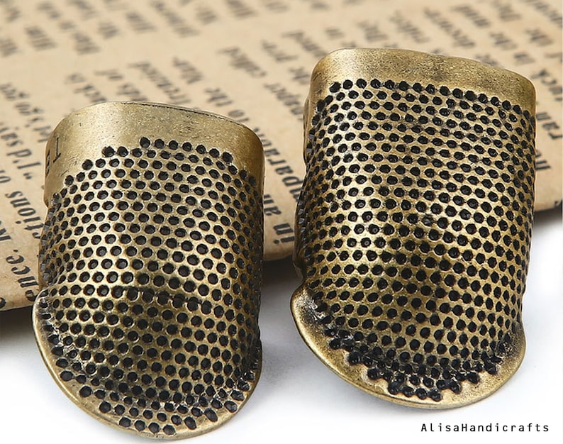 May include: Two antique gold-toned thimbles with a textured, dotted surface. The thimbles are made of metal and have a slightly rounded shape.