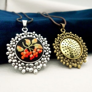 Puede incluir: Dos collares colgantes de plata y oro con un diseño floral. El colgante de plata presenta un fondo negro con un diseño bordado de bayas rojas y hojas verdes. El colgante de oro presenta un disco metálico redondo perforado.