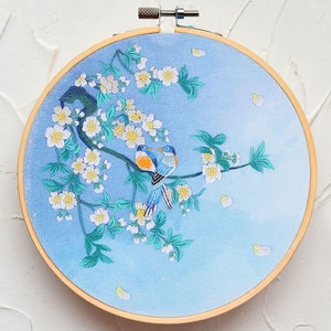 May include: Embroidered hoop art featuring two blue birds perched on a branch with white blossoms. The background is a light blue gradient.