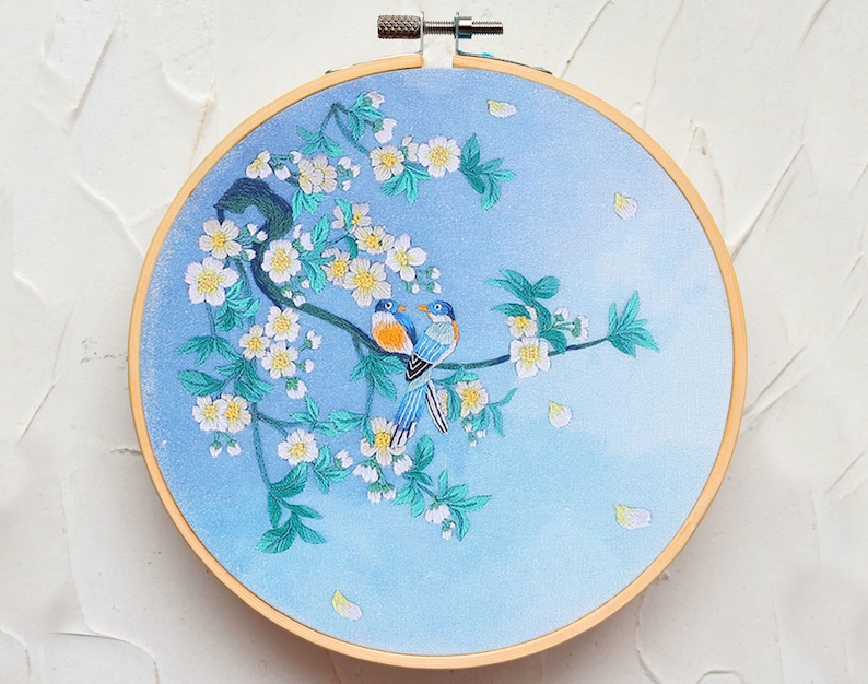 May include: Embroidered hoop art featuring two blue birds perched on a branch with white blossoms. The background is a light blue gradient.