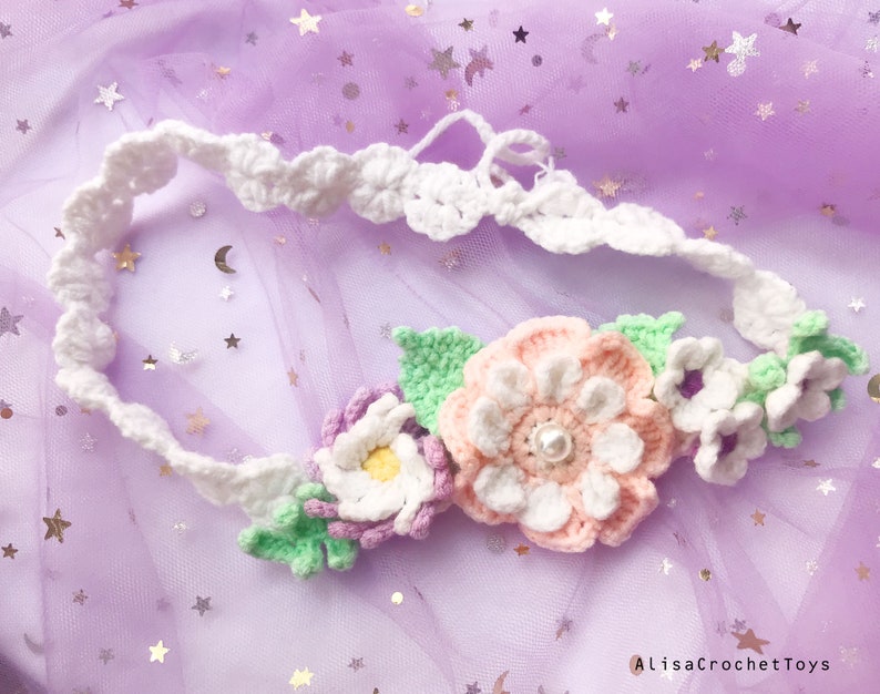 May include: A white crocheted headband with a floral design. The flowers are pink, white, and green. The headband is on a purple background.