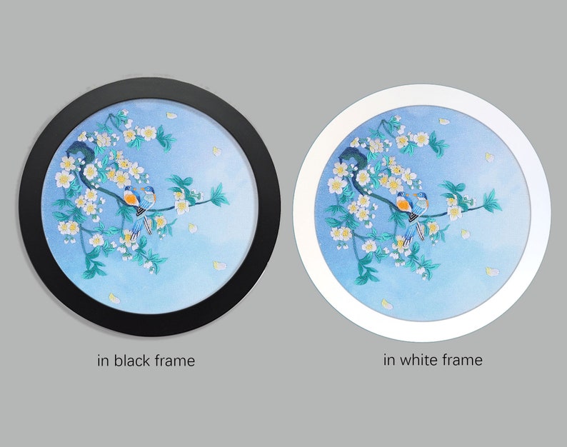 May include: A round embroidery depicting two blue birds perched on a branch with white blossoms against a blue watercolor background. The embroidery is framed in black and white.