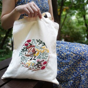 Puede incluir: Una bolsa de lona blanca con un dise&ntilde;o de bordado floral y de p&aacute;jaro. El bordado presenta un p&aacute;jaro peque&ntilde;o, flores y hojas en tonos rojos, amarillos y verdes.