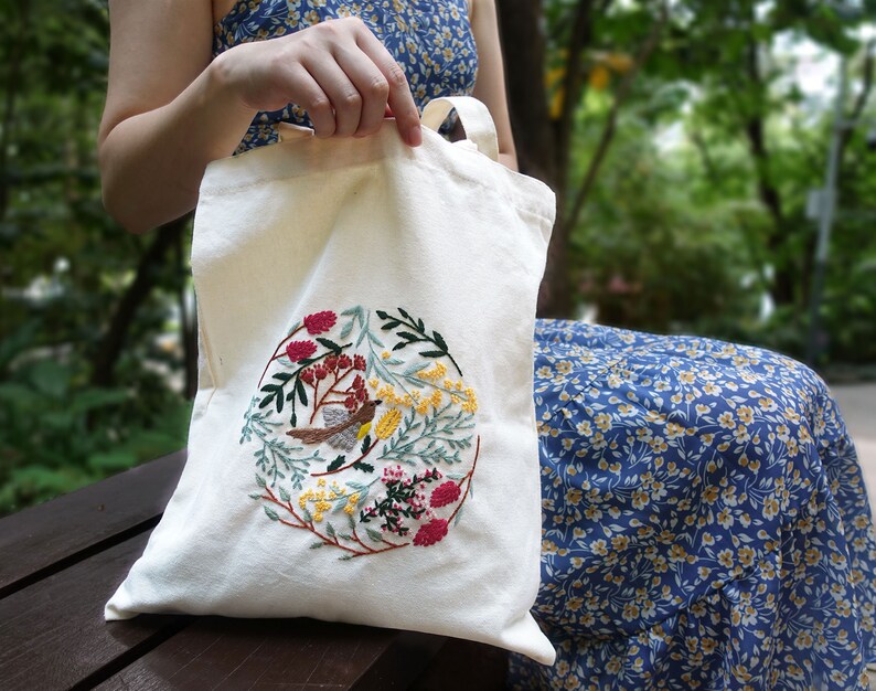 Puede incluir: Una bolsa de lona blanca con un dise&ntilde;o de bordado floral y de p&aacute;jaro. El bordado presenta un p&aacute;jaro peque&ntilde;o, flores y hojas en tonos rojos, amarillos y verdes.