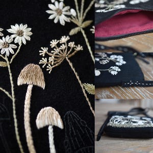 May include: A black crossbody bag featuring detailed embroidery of white daisies and tan mushrooms. The bag has a dark pink interior lining, a black zipper closure, and a thin black strap with white decorative stitching along the edges.