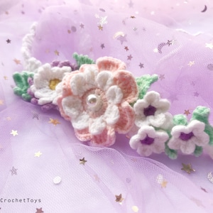 May include: A crocheted flower crown with white, pink, and purple flowers and green leaves. The crown is on a purple background with silver glitter stars.