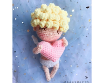 May include: A crocheted amigurumi cupid doll with yellow hair, pink cheeks, and closed eyes. The doll is holding a pink heart in its hands and is wearing a white diaper. The doll is lying on a blue background with white stars.