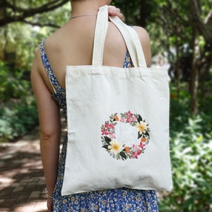 Puede incluir: Una bolsa de lona blanca con una corona de flores bordada en la parte delantera. La corona est&aacute; hecha de flores rosas, amarillas y naranjas con hojas verdes.