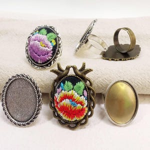 May include: Five silver and brass ring blanks with different designs. Two blanks feature a floral embroidery design, one with a purple flower and the other with a red and yellow flower. The other three blanks are plain, one with a silver finish and two with a brass finish.