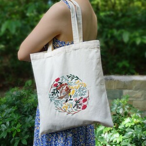 Puede incluir: Una bolsa de lona blanca con un dise&ntilde;o bordado circular que presenta un p&aacute;jaro, flores y hojas en tonos rojos, amarillos, verdes y azules.
