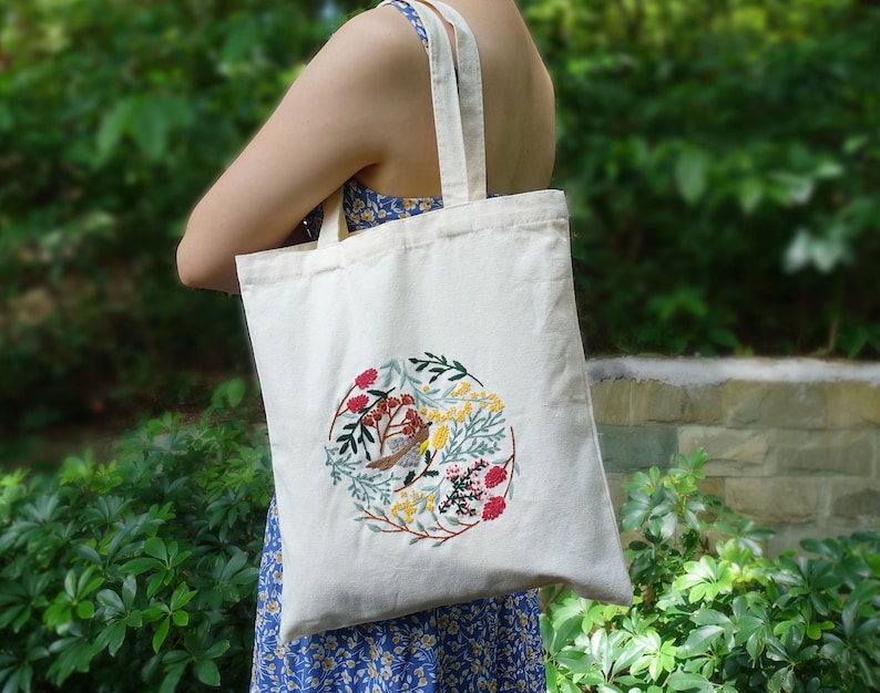 Puede incluir: Una bolsa de lona blanca con un dise&ntilde;o bordado circular que presenta un p&aacute;jaro, flores y hojas en tonos rojos, amarillos, verdes y azules.