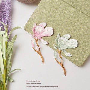 May include: Two embroidered fabric patches featuring pink and green magnolia flowers with brown stems. The patches are on a green fabric background.