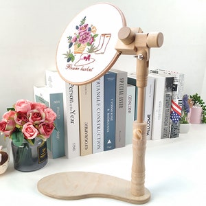 May include: A wooden embroidery hoop stand with a round base and a wooden arm that can be adjusted to different angles. The stand is holding a white embroidery hoop with a floral design and the text "Flower basket".