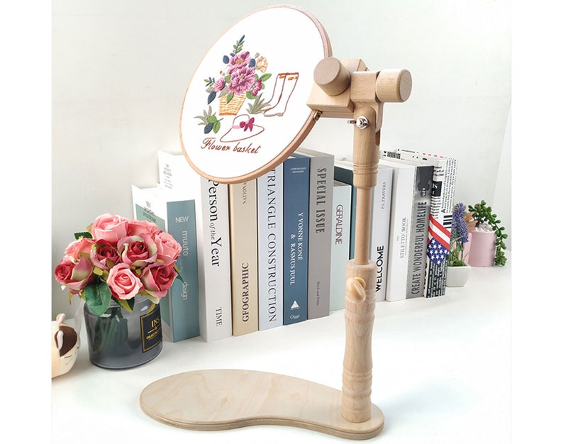 May include: A wooden embroidery hoop stand with a round base and a wooden arm that can be adjusted to different angles. The stand is holding a white embroidery hoop with a floral design and the text "Flower basket".