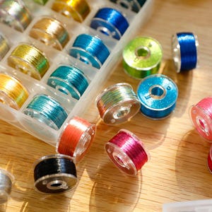 Puede incluir: Una colección de coloridas bobinas de máquina de coser, incluyendo plateadas, doradas, turquesas, azules y rosas, están dispuestas en un organizador de plástico transparente y dispersas sobre una superficie de madera. Las bobinas están llenas de hilo.