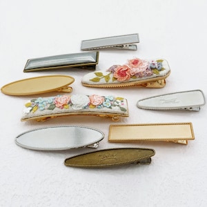 May include: A collection of hair clips including two embroidered clips with pink and blue floral patterns on white fabric, several metallic clips in gold, silver, and bronze tones with smooth or textured surfaces, and one clip featuring the text Denisa.