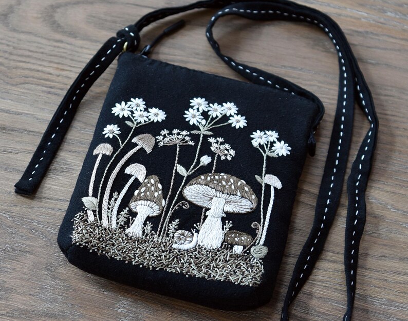 May include: A black crossbody bag featuring detailed white and brown embroidery of various mushrooms, small flowers, and a snail on the front. The bag has a zipper closure and a long black strap with white decorative stitching along the edges.