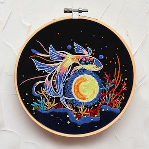 May include: Embroidered artwork of a colorful fish swimming near a yellow and orange orb, possibly a sun or moon, against a black background. The fish has blue, yellow, and pink details. The artwork is framed in a wooden embroidery hoop.