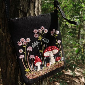 May include: A black crossbody bag featuring detailed embroidery of red and white mushrooms alongside pink wildflowers and green stems. The bag has a zipper closure at the top and a black strap, with the design centered on the front fabric panel.