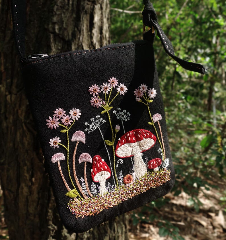 May include: A black crossbody bag featuring detailed embroidery of red and white mushrooms alongside pink wildflowers and green stems. The bag has a zipper closure at the top and a black strap, with the design centered on the front fabric panel.