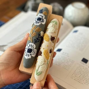 Könnte beinhalten: Zwei hölzerne Stickrahmen mit Blumendesigns. Der Rahmen links hat weiße Blüten mit schwarzen Zentren und grünen Blättern auf grauem Hintergrund. Der Rahmen rechts hat weiße Blüten mit gelben Zentren und braunen Blättern auf beigefarbenem Hintergrund.