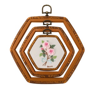 May include: A set of three brown hexagonal embroidery hoops, nested together. The center hoop displays a floral embroidery of pink roses with green leaves and a blue ribbon. The word "Rose" is embroidered below the flowers. The hoops have metal hanging loops.