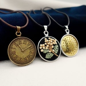 May include: Three necklaces with different pendants. The first pendant is a round, antique-style clock face with Roman numerals. The second pendant is a round, black fabric with a white flower embroidered on it. The third pendant is a round, gold-colored metal with a perforated design.