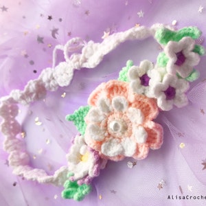 May include: A white crocheted headband with a floral design. The headband is decorated with white, pink, and purple flowers, as well as green leaves.