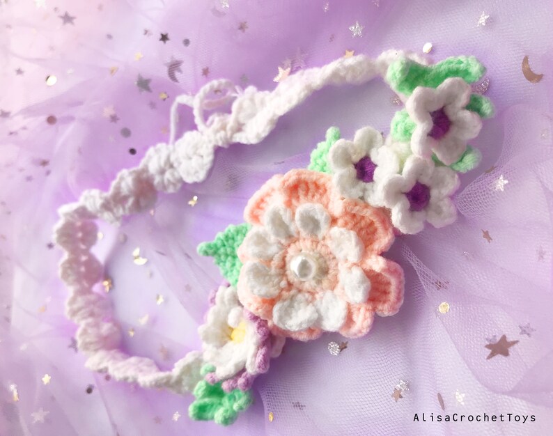 May include: A white crocheted headband with a floral design. The headband is decorated with white, pink, and purple flowers, as well as green leaves.