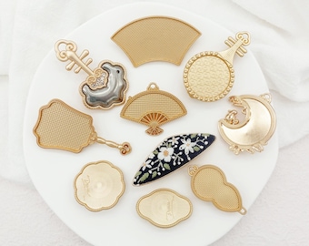 Puede incluir: Una colección de dijes y adornos decorativos dorados en varias formas, incluyendo abanicos, instrumentos musicales y una luna creciente. Un dije es un óvalo negro con bordado floral blanco. Los dijes están dispuestos sobre una superficie blanca.