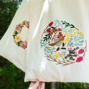 Puede incluir: Una bolsa de lona blanca con dos dise&ntilde;os bordados. Un dise&ntilde;o presenta una corona de flores en tonos de rosa, rojo, amarillo y verde. El otro dise&ntilde;o presenta un p&aacute;jaro rodeado de flores y hojas en tonos de verde, amarillo, rosa y rojo.