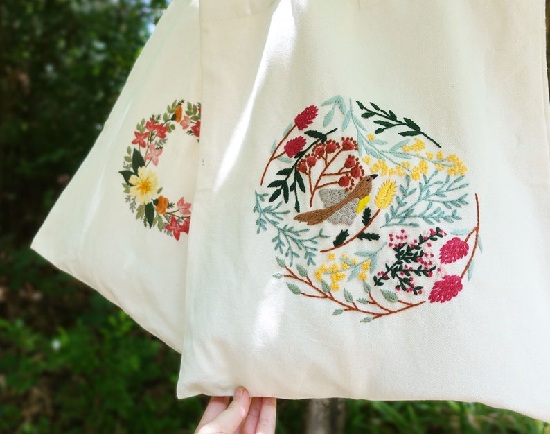 Puede incluir: Una bolsa de lona blanca con dos dise&ntilde;os bordados. Un dise&ntilde;o presenta una corona de flores en tonos de rosa, rojo, amarillo y verde. El otro dise&ntilde;o presenta un p&aacute;jaro rodeado de flores y hojas en tonos de verde, amarillo, rosa y rojo.
