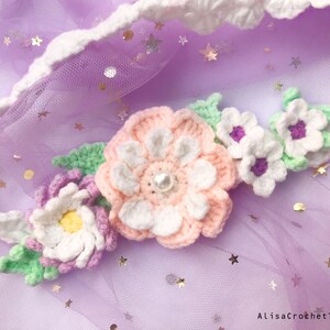 May include: A crocheted flower headband with pink, white, and green flowers. The headband is on a purple background with silver glitter stars.
