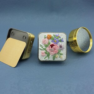 May include: Three decorative items: a small, open, rectangular brass box with a gold interior, a square embroidered patch with a floral design, and a gold-colored, round object with a textured surface.