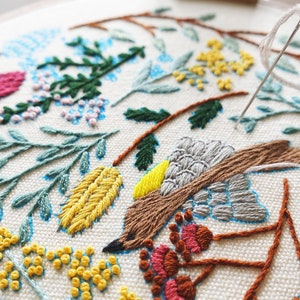 Puede incluir: Primer plano de un dise&ntilde;o de bordado hecho a mano que presenta un p&aacute;jaro marr&oacute;n con acentos grises y amarillos, rodeado de motivos florales verdes, azules y amarillos sobre una tela blanca.