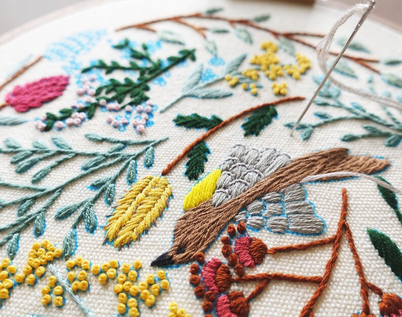 Puede incluir: Primer plano de un dise&ntilde;o de bordado hecho a mano que presenta un p&aacute;jaro marr&oacute;n con acentos grises y amarillos, rodeado de motivos florales verdes, azules y amarillos sobre una tela blanca.