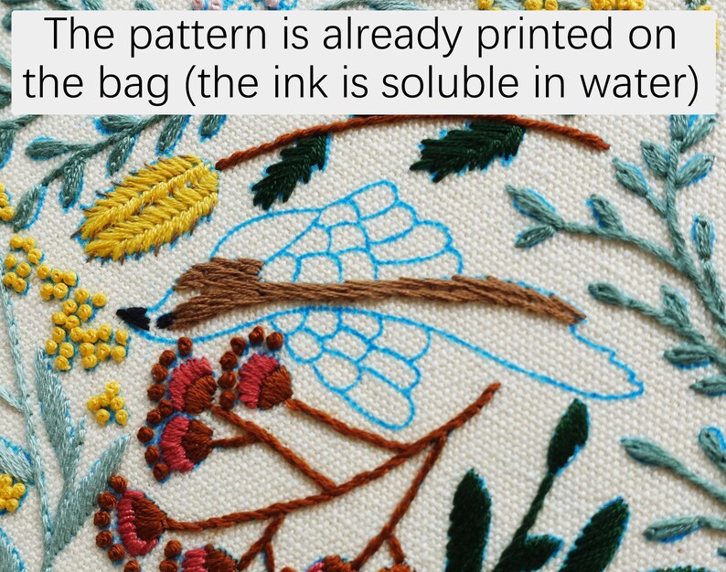 Puede incluir: Primer plano de una tela blanca con un p&aacute;jaro azul, una rama marr&oacute;n y hojas verdes y amarillas bordadas sobre ella. El texto "The pattern is already printed on the bag (the ink is soluble in water)" es visible en la parte superior de la imagen.